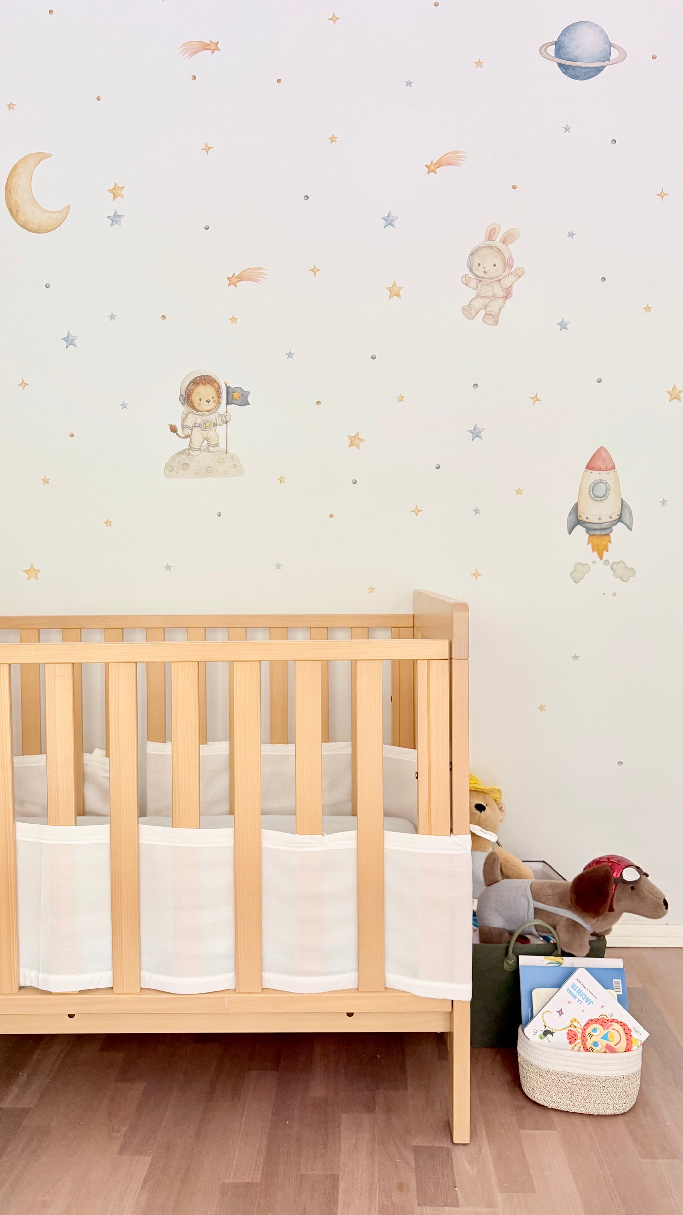 White nursery wall decorated with Lemon and Bubbles’ Space and Beyond fabric wall decals: a lion and bunny astronaut, rocket, moon, ringed planet, shooting stars, and gold and silver stars. Below, a light wood cot with a white skirt, and a basket with stuffed animals and books on the floor.