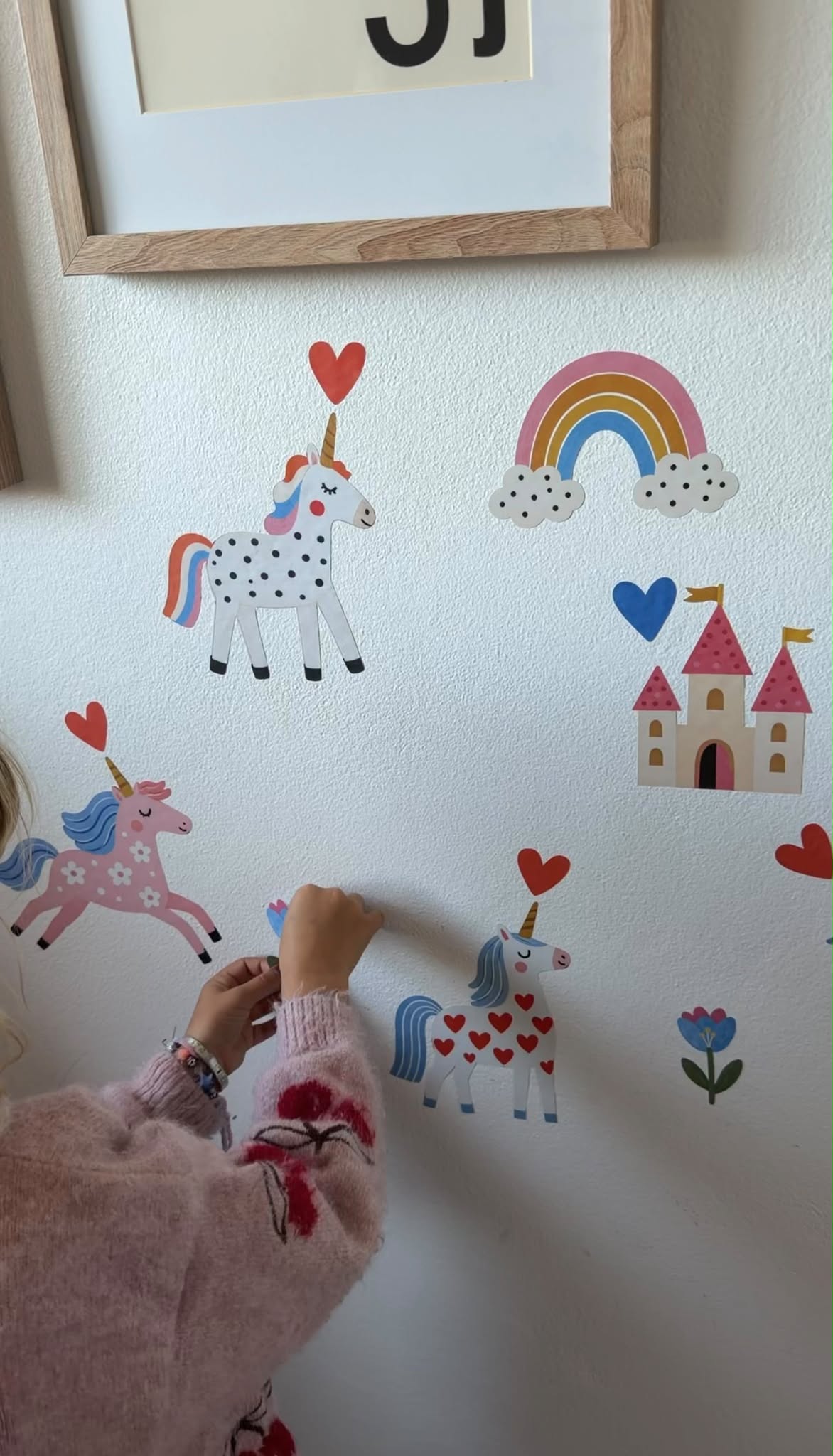 Child placing a Twinkle Kingdom wall sticker among colourful unicorns, a rainbow, hearts and a castle on a bedroom wall.